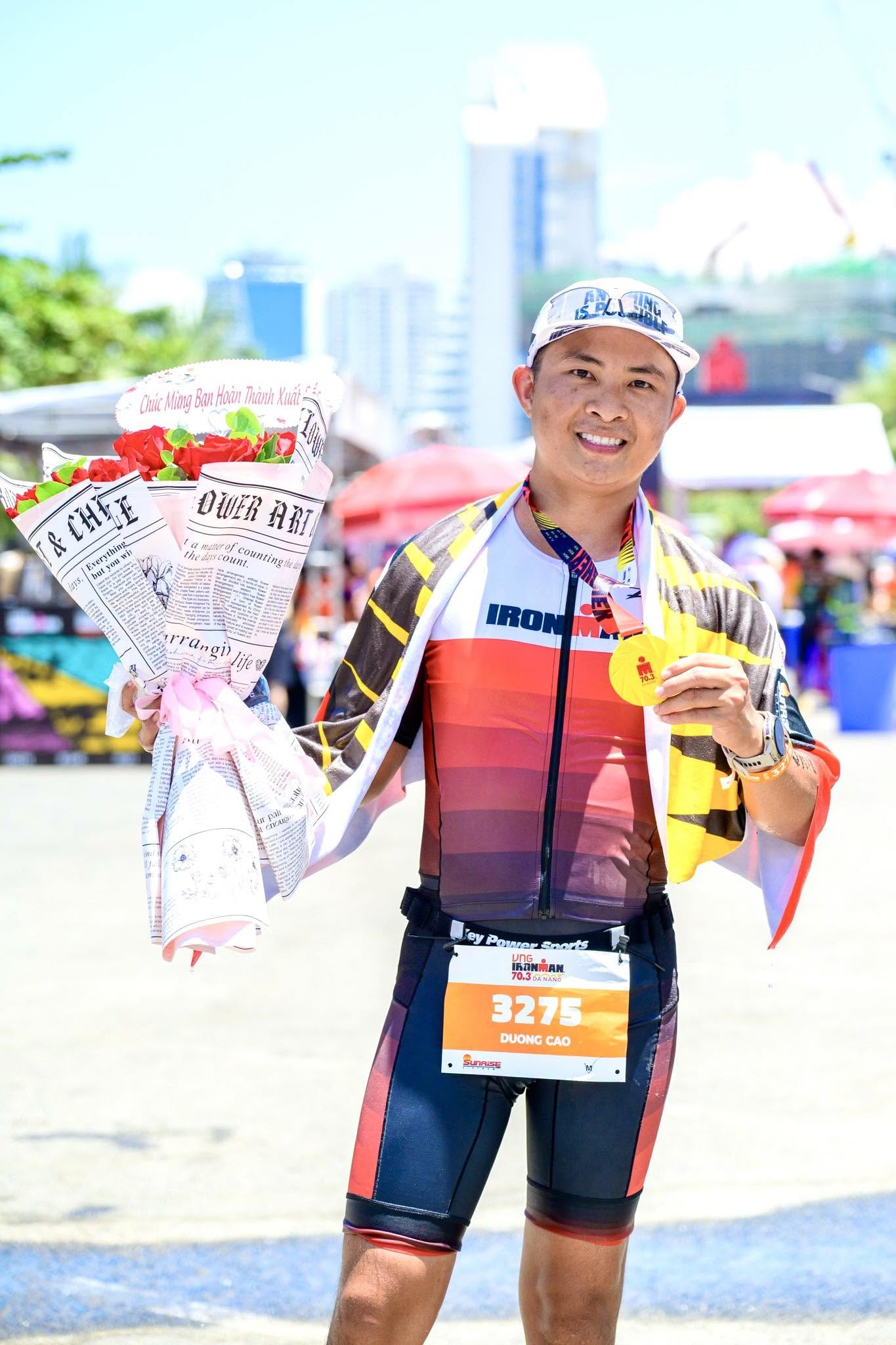 Dương Cao Trí cười rạng rỡ, tay cầm huy chương và bó hoa sau khi hoàn thành Ironman 70.3 Đà Nẵng